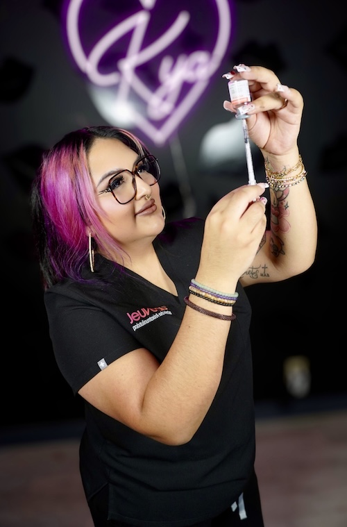 Smiling woman with half pink hair holding beauty product boxes in front of a gray wall.