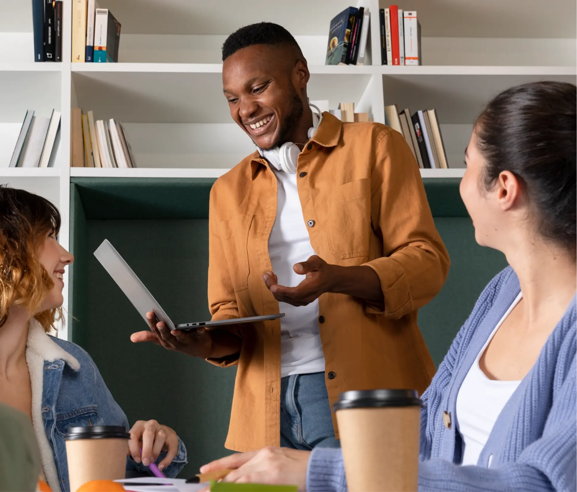 Smiling man showing a laptop to two women seated at a table with coffee cups and books in a casual meeting.