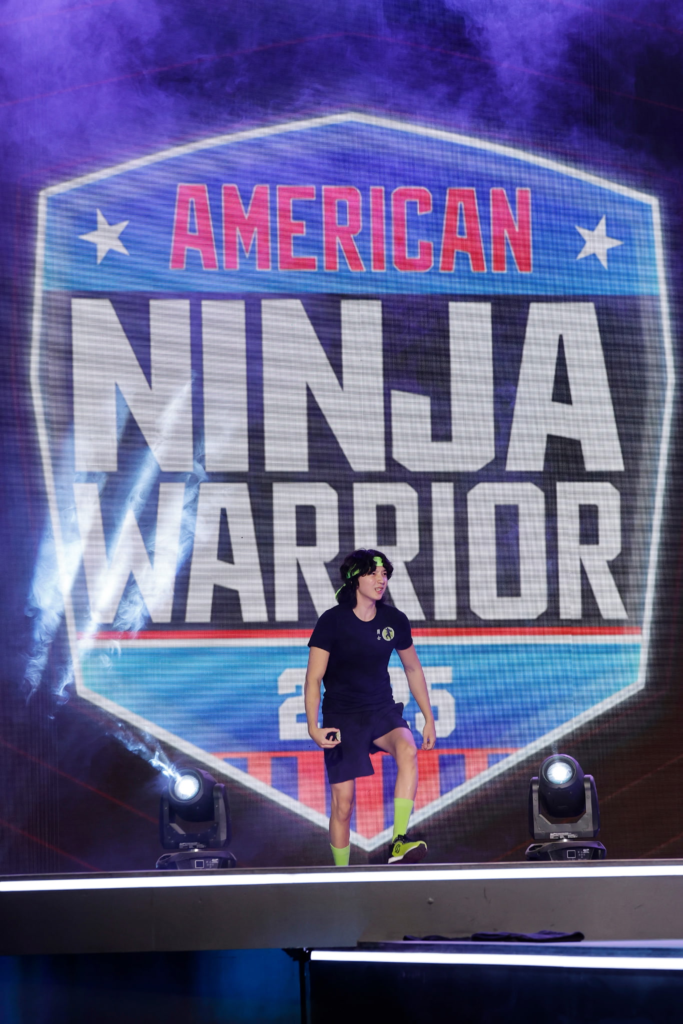 Athlete walking onto the American Ninja Warrior stage under bright lights and competition backdrop