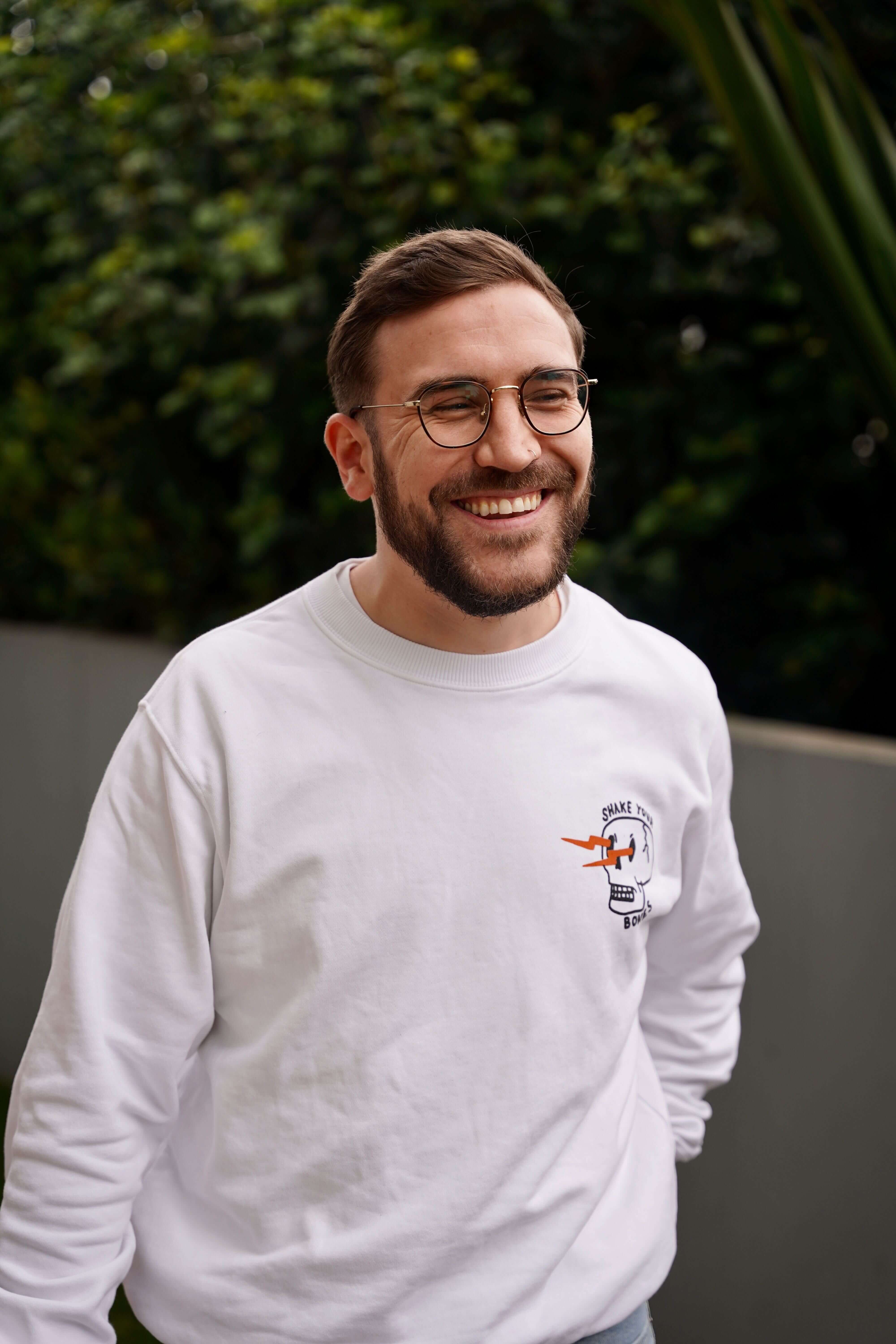 Smiling man with glasses and beard wearing a white sweatshirt with a skull and lightning design standing outdoors.