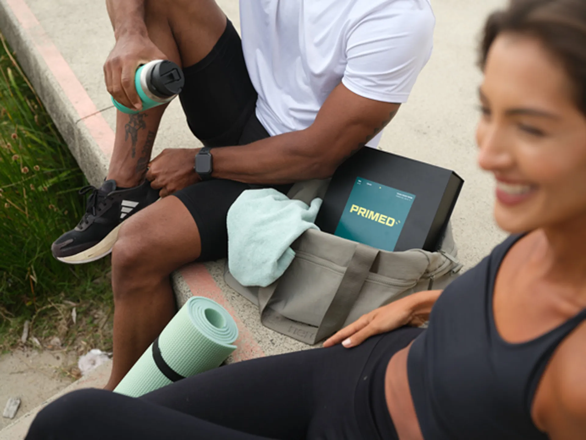 Man and woman in workout clothes sitting outdoors with exercise mat and a bag containing a box labeled PRIMED.