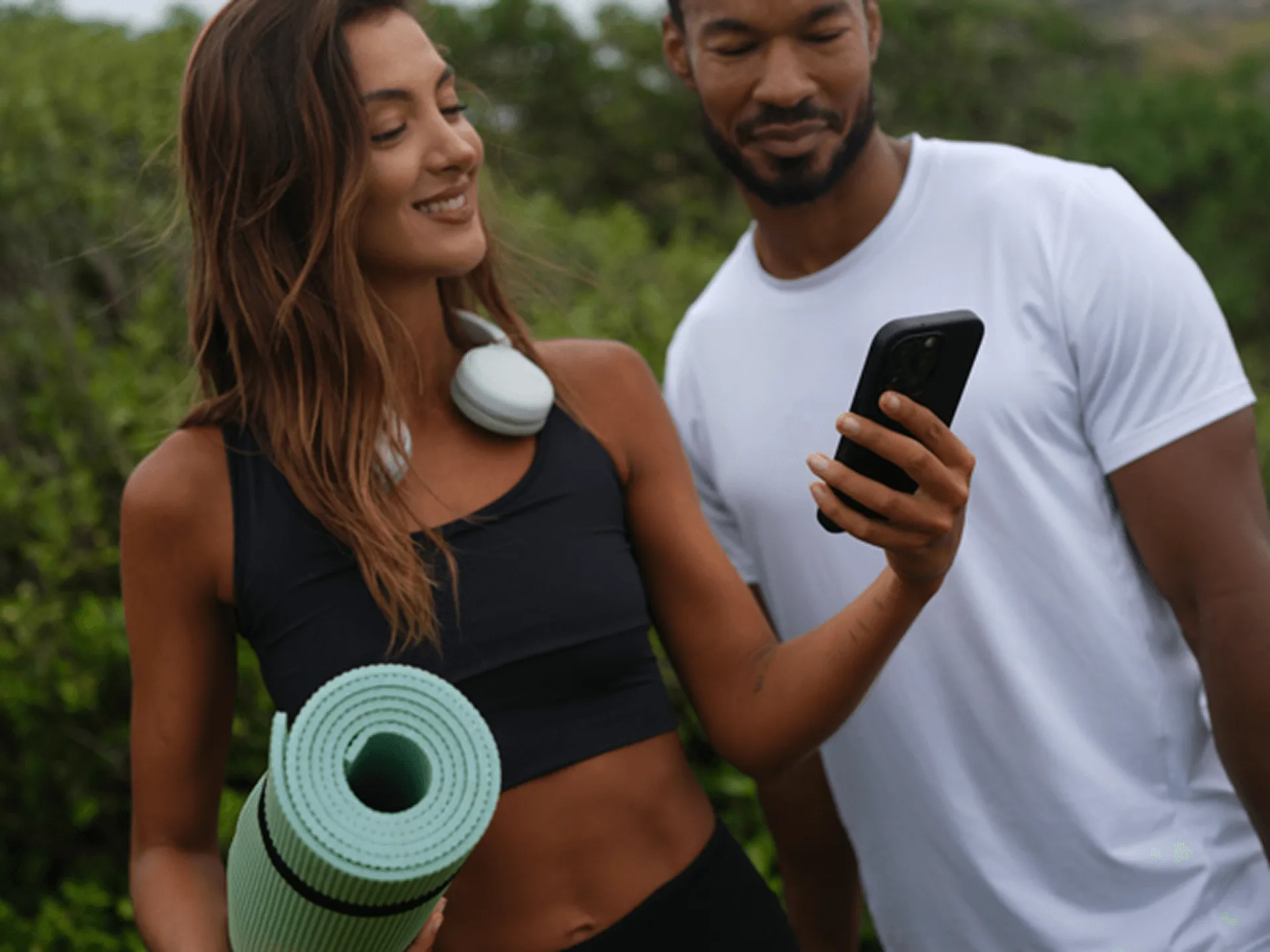 Smiling woman holding a rolled yoga mat and wearing headphones around her neck, standing next to a man in a white t-shirt looking at a phone outdoors.