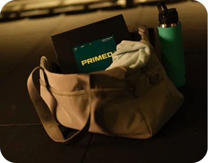 Beige gym bag containing a white towel and a black folder with the word 'PRIMED' on it, next to a green water bottle.