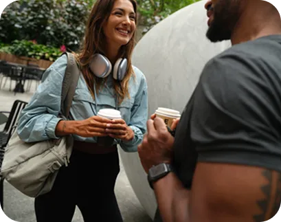 Smiling woman with headphones and tote bag holding a coffee cup, talking to a man with a tattoo and smartwatch outdoors.