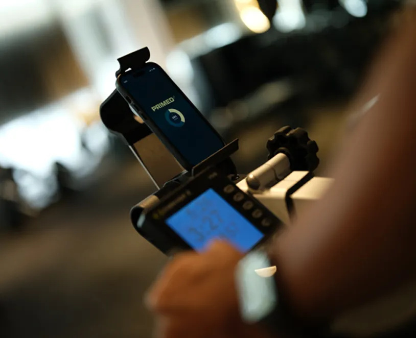 Person operating a stationary exercise bike with a smartphone displaying a fitness app and the bike's control panel showing workout data.