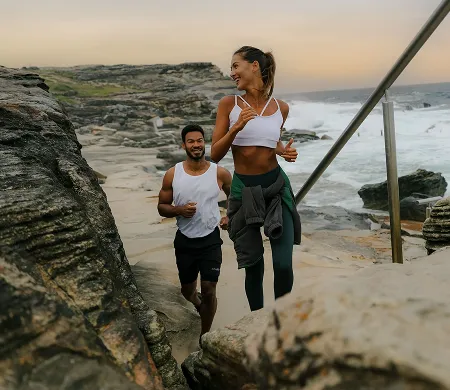 A woman and man jogging together along a rocky coastline during sunset.