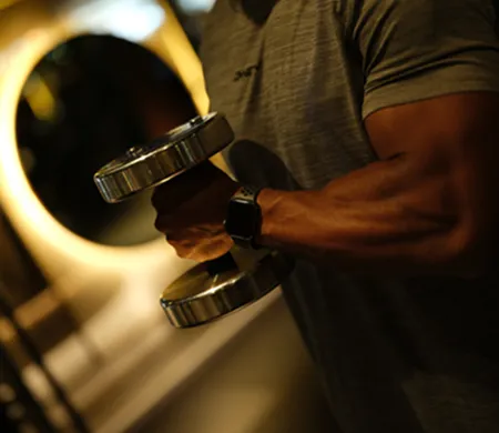Close-up of a muscular arm lifting a shiny metal dumbbell indoors with a blurred circular light in the background.