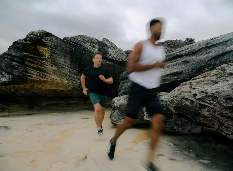 Two men running on a sandy trail near large rock formations under a cloudy sky, with motion blur on the closest runner.