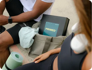 Two people sitting with a gym bag containing a PRIMED box and a yoga mat, one person wearing headphones.