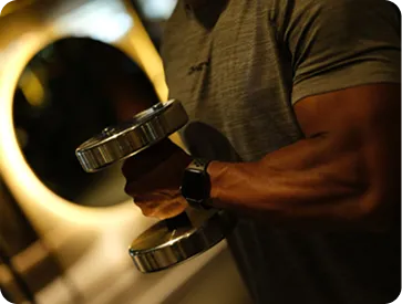 Person lifting a silver dumbbell with muscular arm in dimly lit environment.