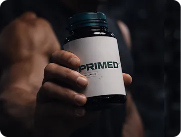 Muscular person holding a dark bottle labeled 'PRIMED' with a white label, in a dimly lit setting.