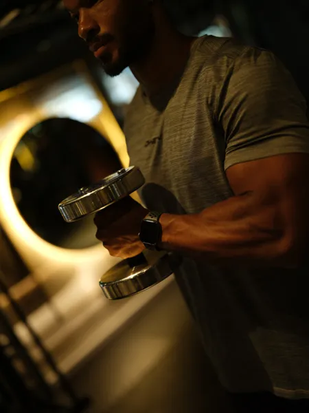 Muscular man in gray t-shirt lifting a silver dumbbell in a dimly lit gym.