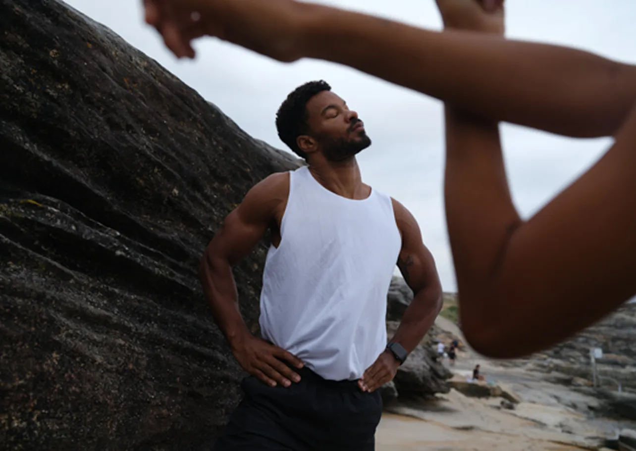 Man in a white sleeveless shirt stretching with eyes closed near rocky beach.