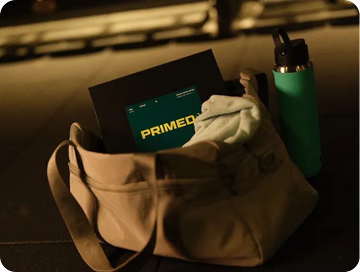 Beige gym bag containing a towel and a booklet labeled 'PRIMED,' next to a green water bottle on a dark surface.