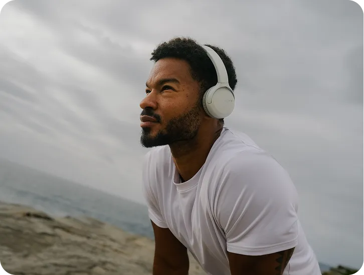 Man wearing white headphones and a white t-shirt looking thoughtfully into the distance by the rocky shore under a cloudy sky.