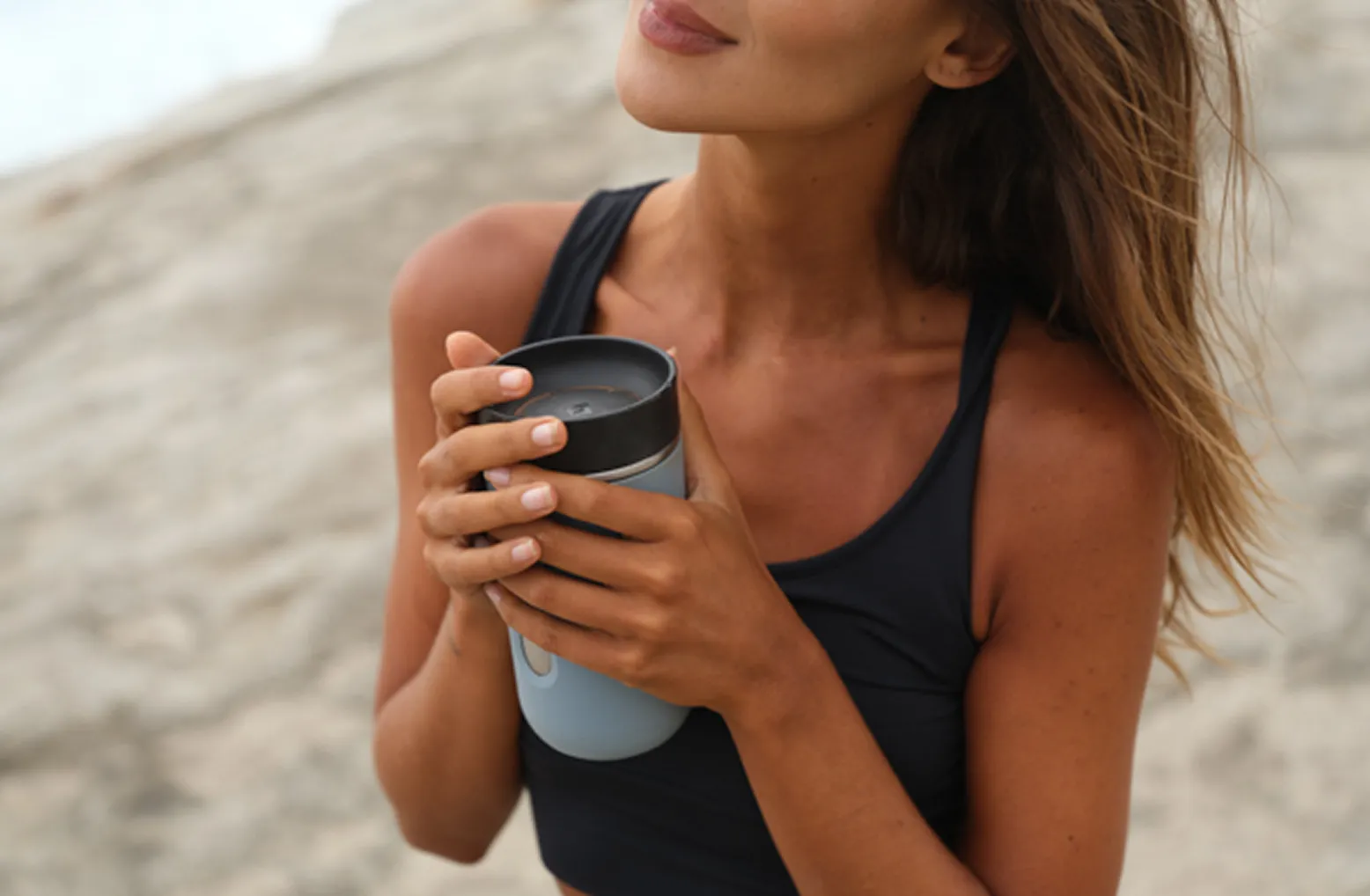 Woman in black sportswear holding a blue travel mug near her chest at the beach.