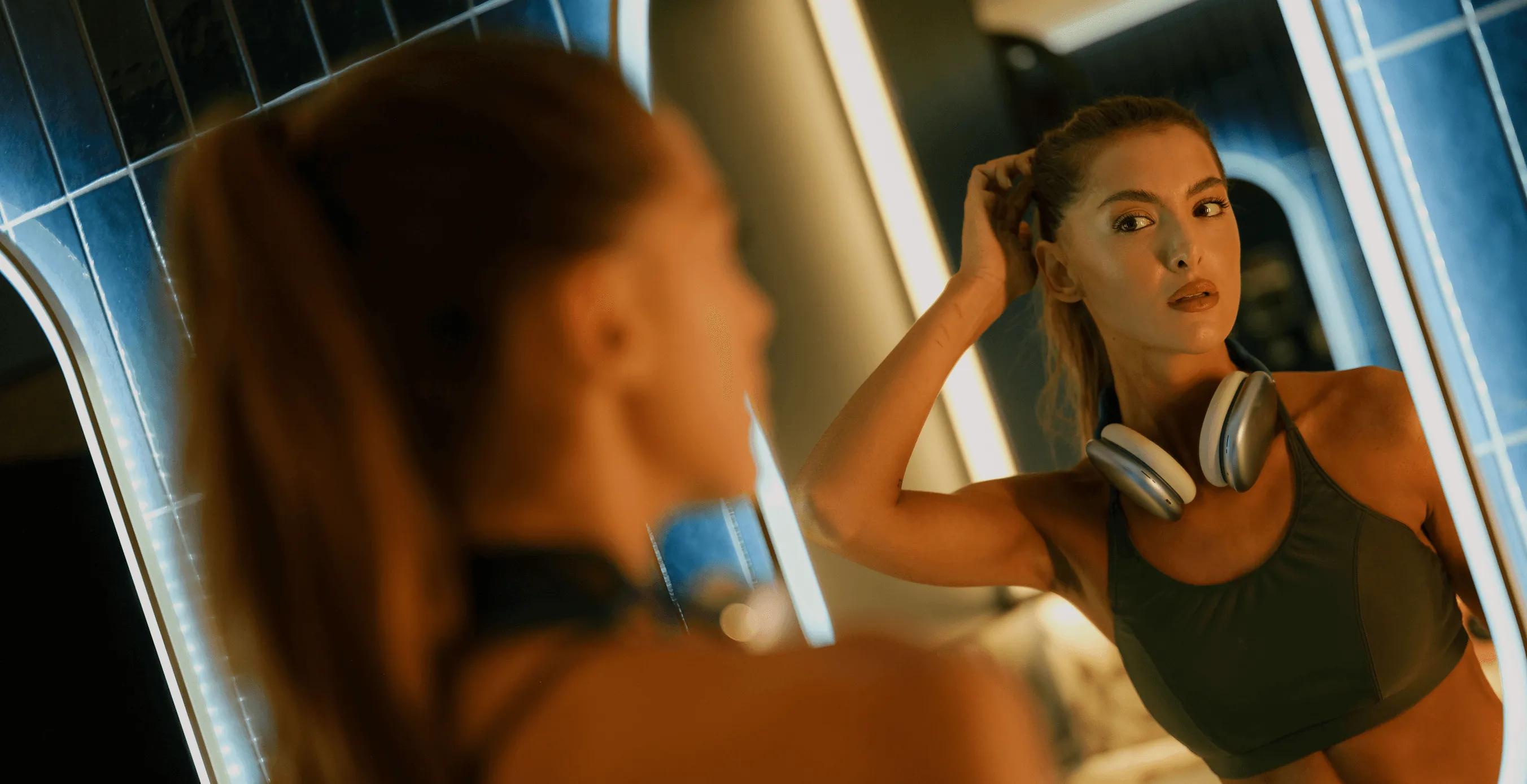 Young woman in workout attire with headphones around her neck, looking at her reflection in a lighted mirror.
