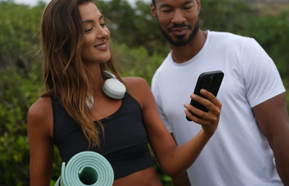Smiling woman with headphones around her neck holding a yoga mat and phone, standing next to a man in a white shirt outdoors.