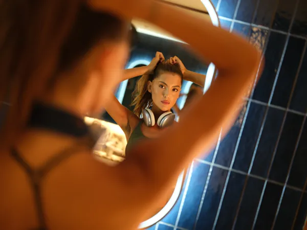 Young woman with headphones around her neck adjusting her hair while looking into a bathroom mirror with blue tiled walls.