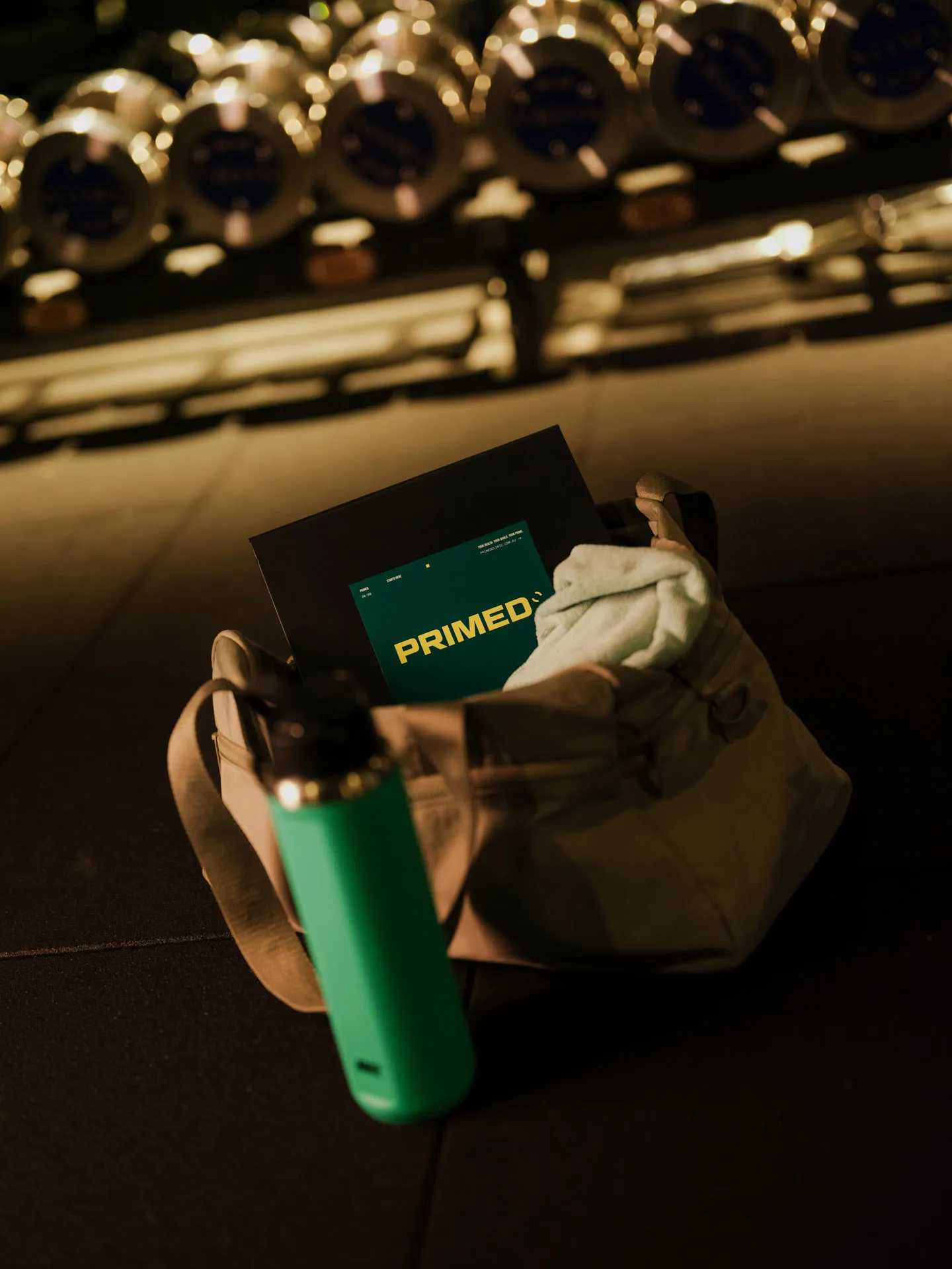 A beige gym bag on the floor with a green water bottle in front and a black folder labeled 'PRIMED' inside the bag.
