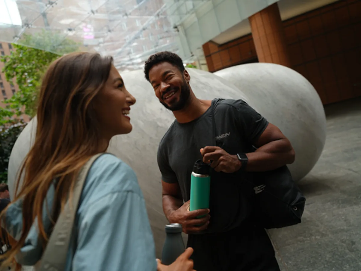 Smiling man and woman holding water bottles chatting outdoors near a large sculpture.