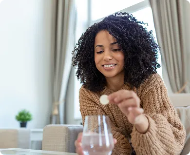Smiling woman in a brown sweater holding a pill and glass of water sitting on a couch.