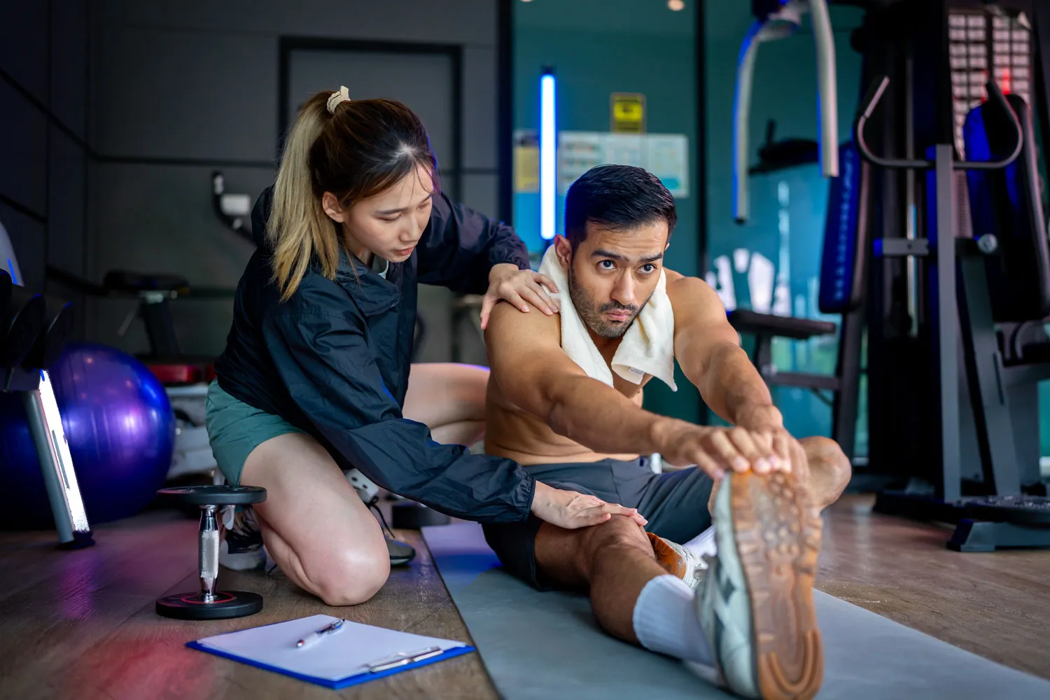 Male athlete stretching forward on a mat while a female trainer assists him in a gym.