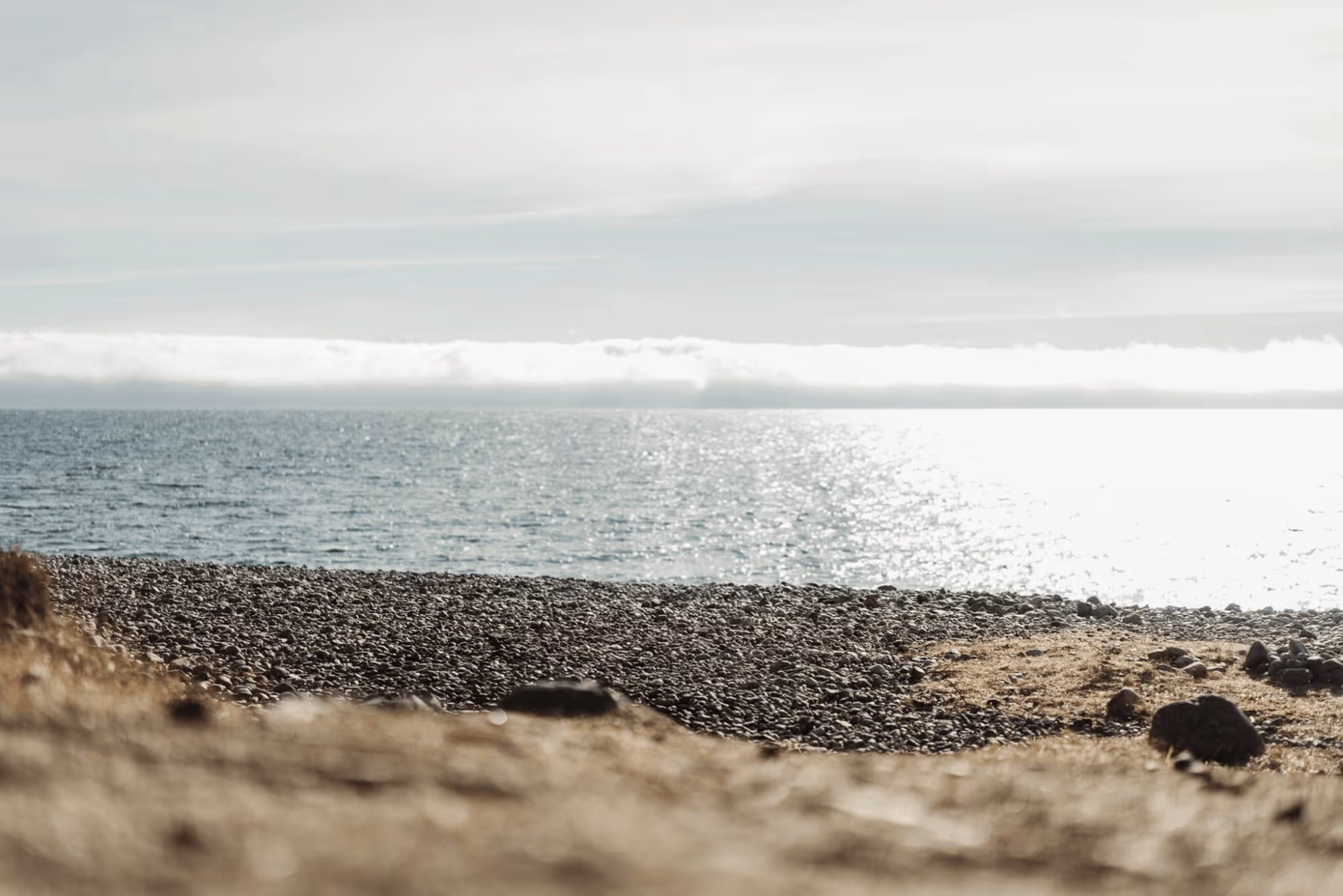 Pebbled beach with calm sea and cloudy sky reflecting sunlight on the water.