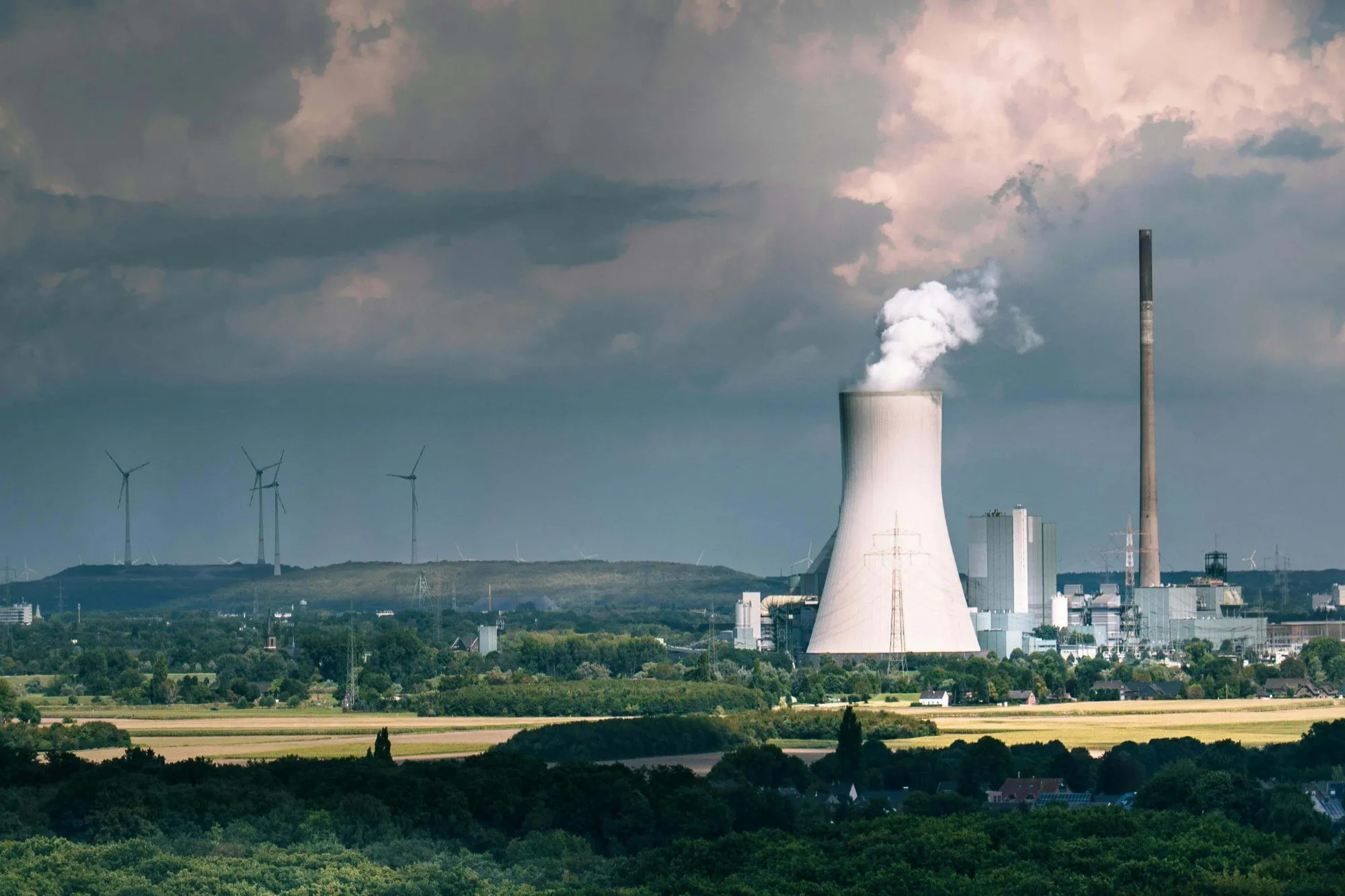 Power plant with cooling tower emitting steam, surrounded by green fields and wind turbines in the background under a cloudy sky.