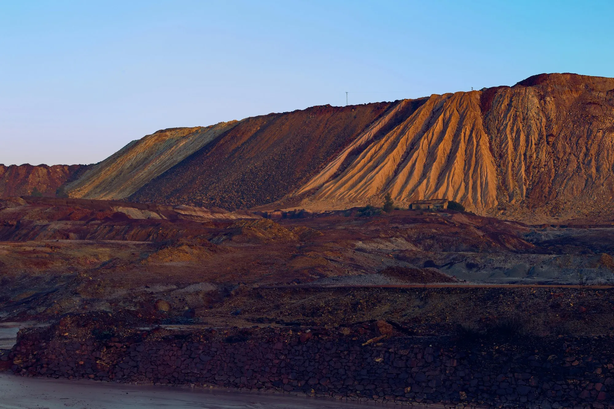Large open-pit mine with terraced slopes under a clear blue sky during sunset.