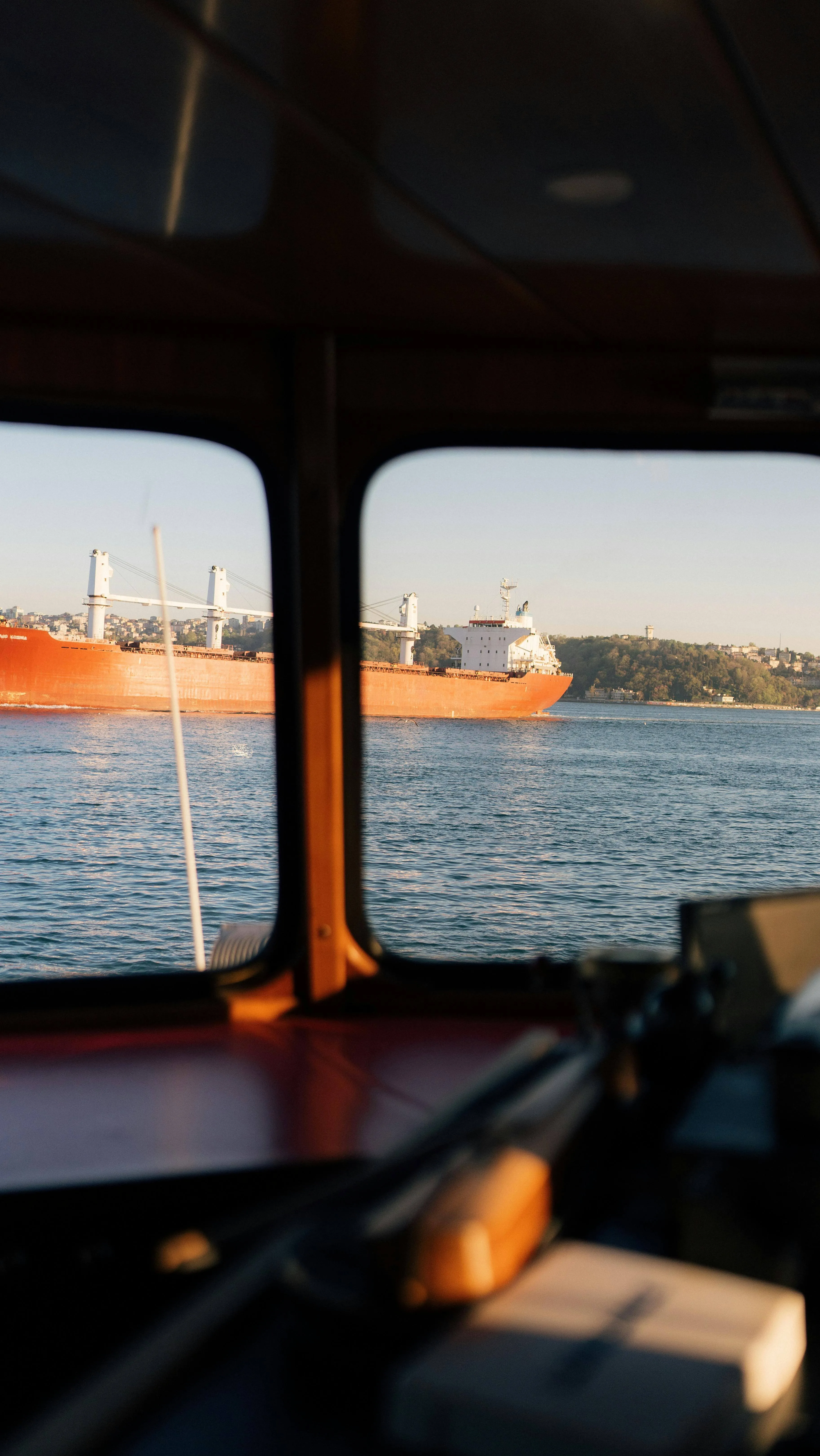 Large orange cargo ship on water viewed through boat window with blurred interior foreground.