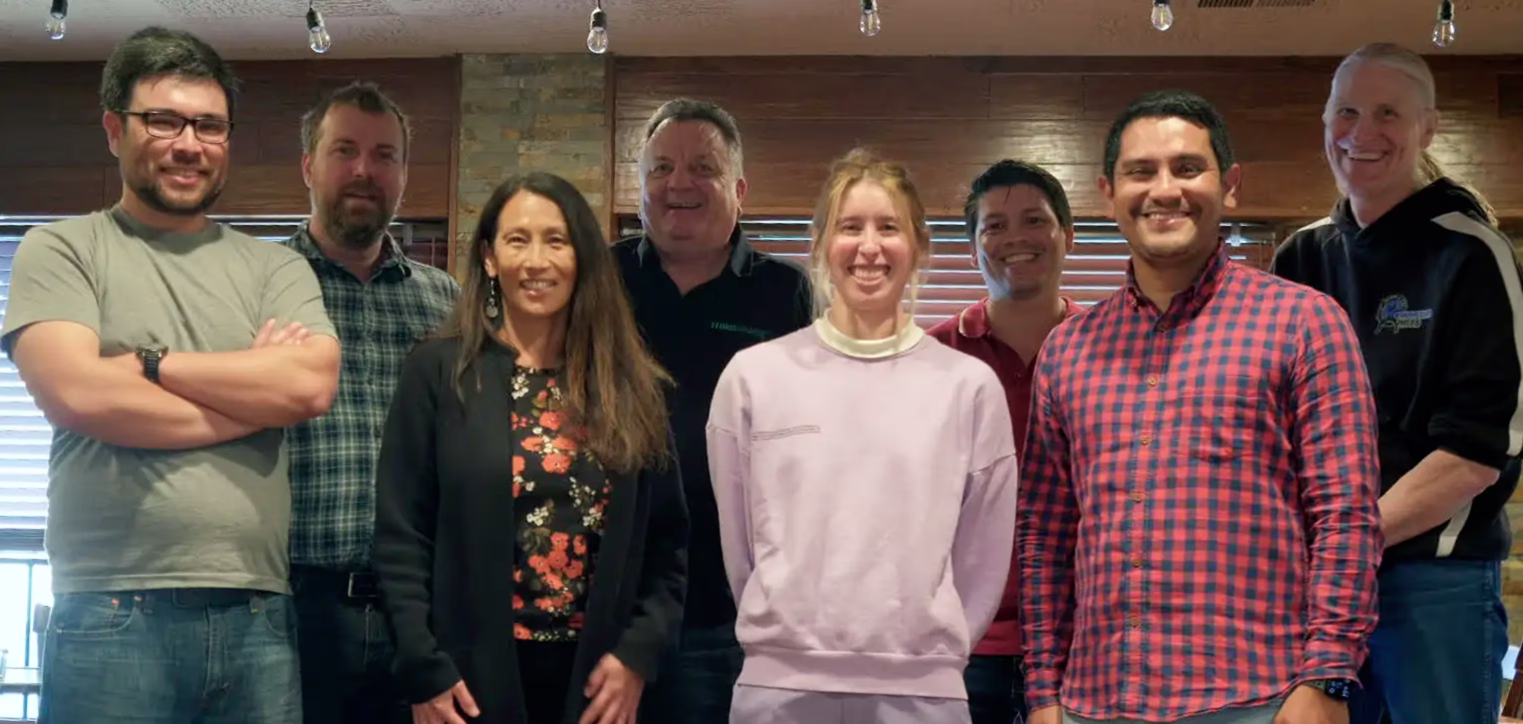 Group photo of eight smiling people standing indoors with wooden walls and hanging lightbulbs above.