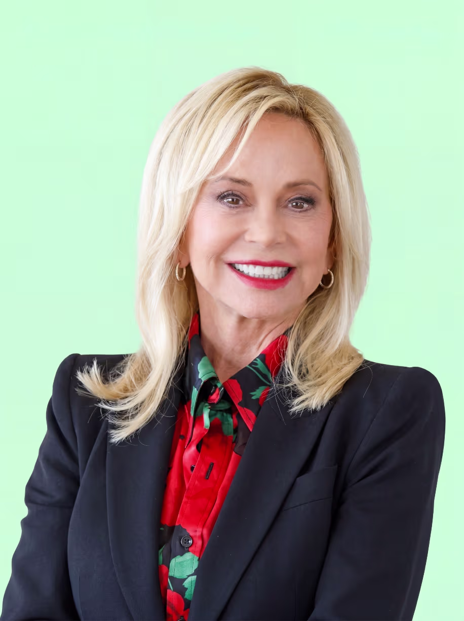 Smiling blonde woman wearing hoop earrings, black blazer, and red floral blouse against a light green background.