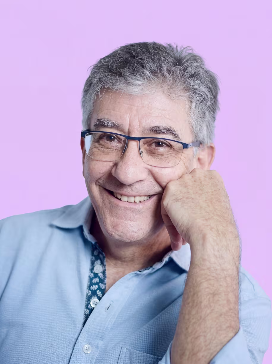 Smiling older man with gray hair and glasses resting his chin on his hand wearing a light blue shirt.