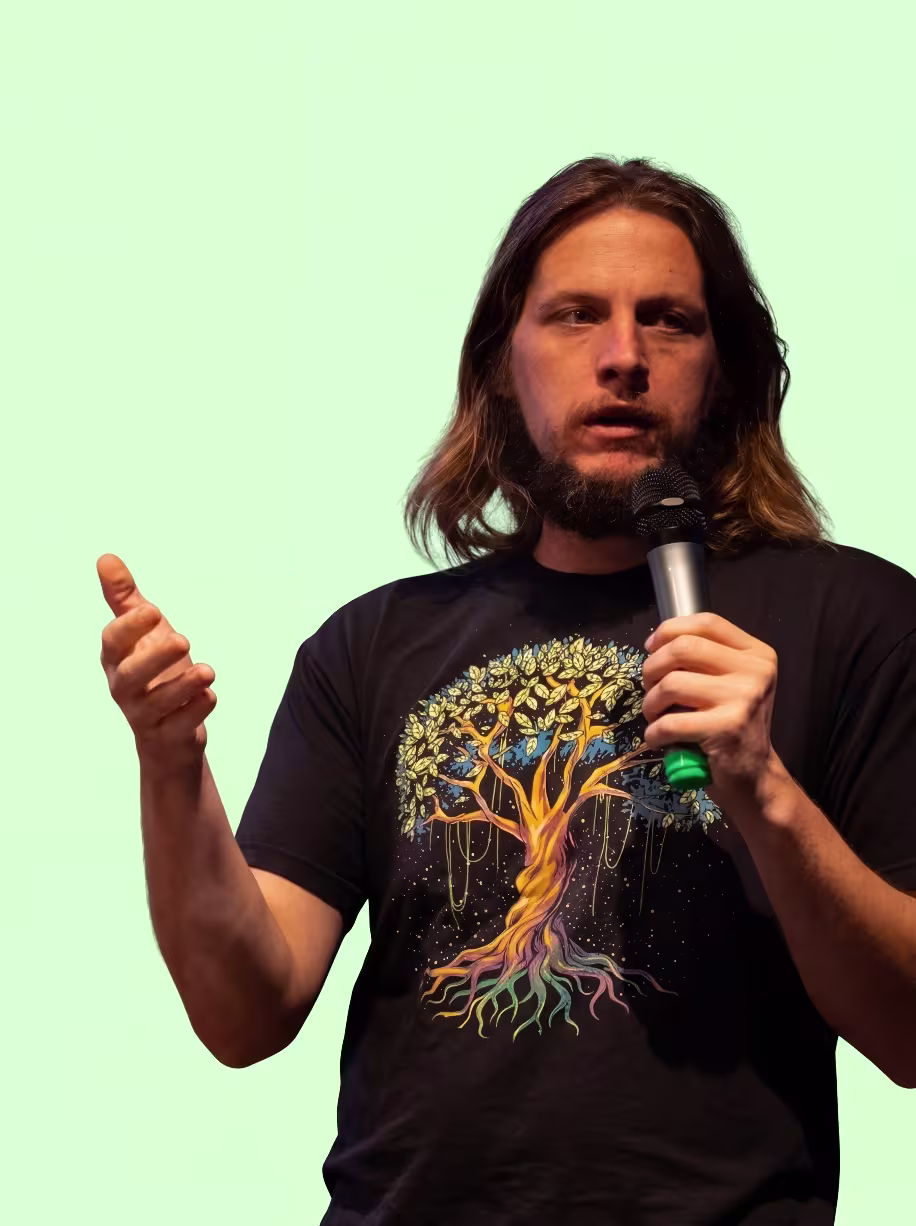 Man with shoulder-length hair speaking into a microphone, wearing a black T-shirt with a colorful tree graphic.