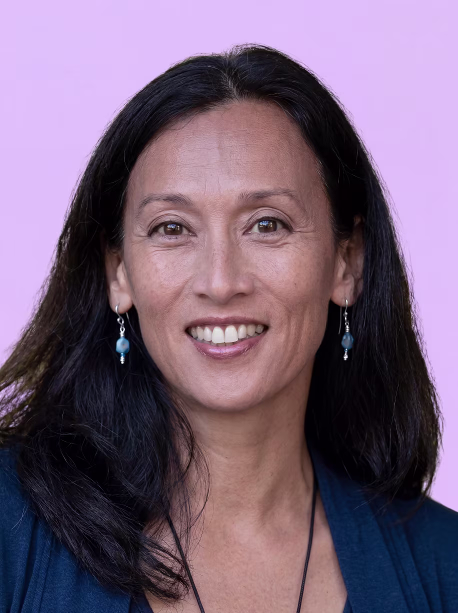 Smiling woman with long dark hair wearing blue earrings and a navy top against a light purple background.