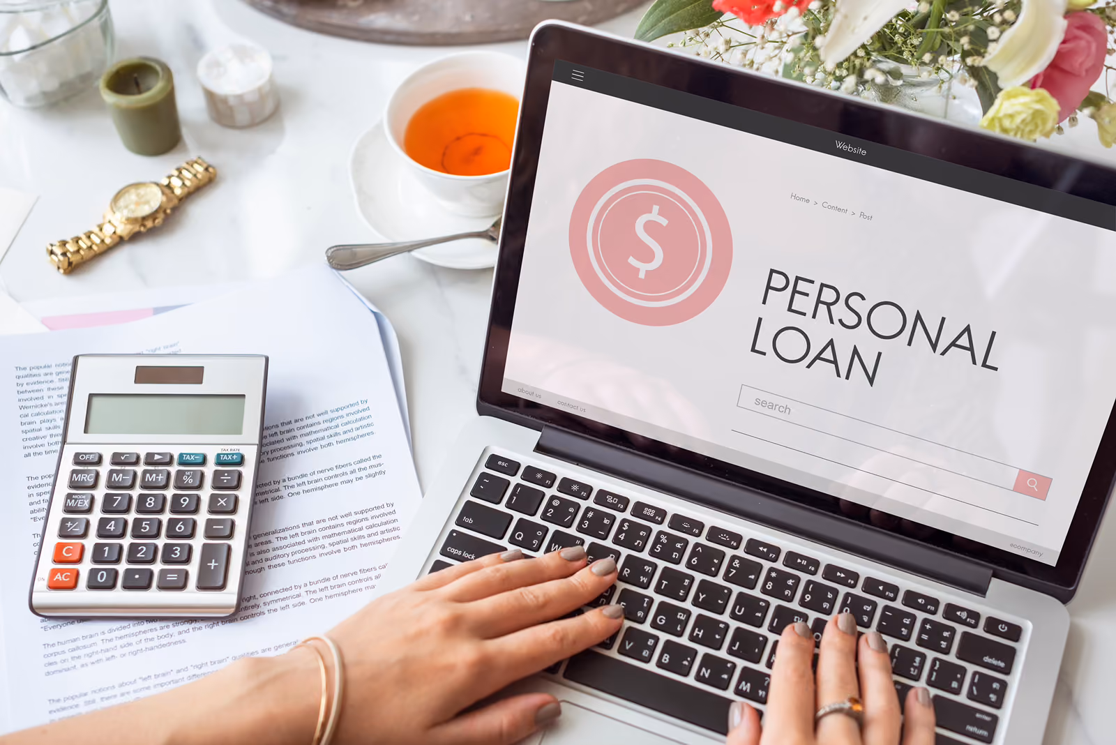 person using a laptop with a personal loan page and a calculator on paperwork to the left