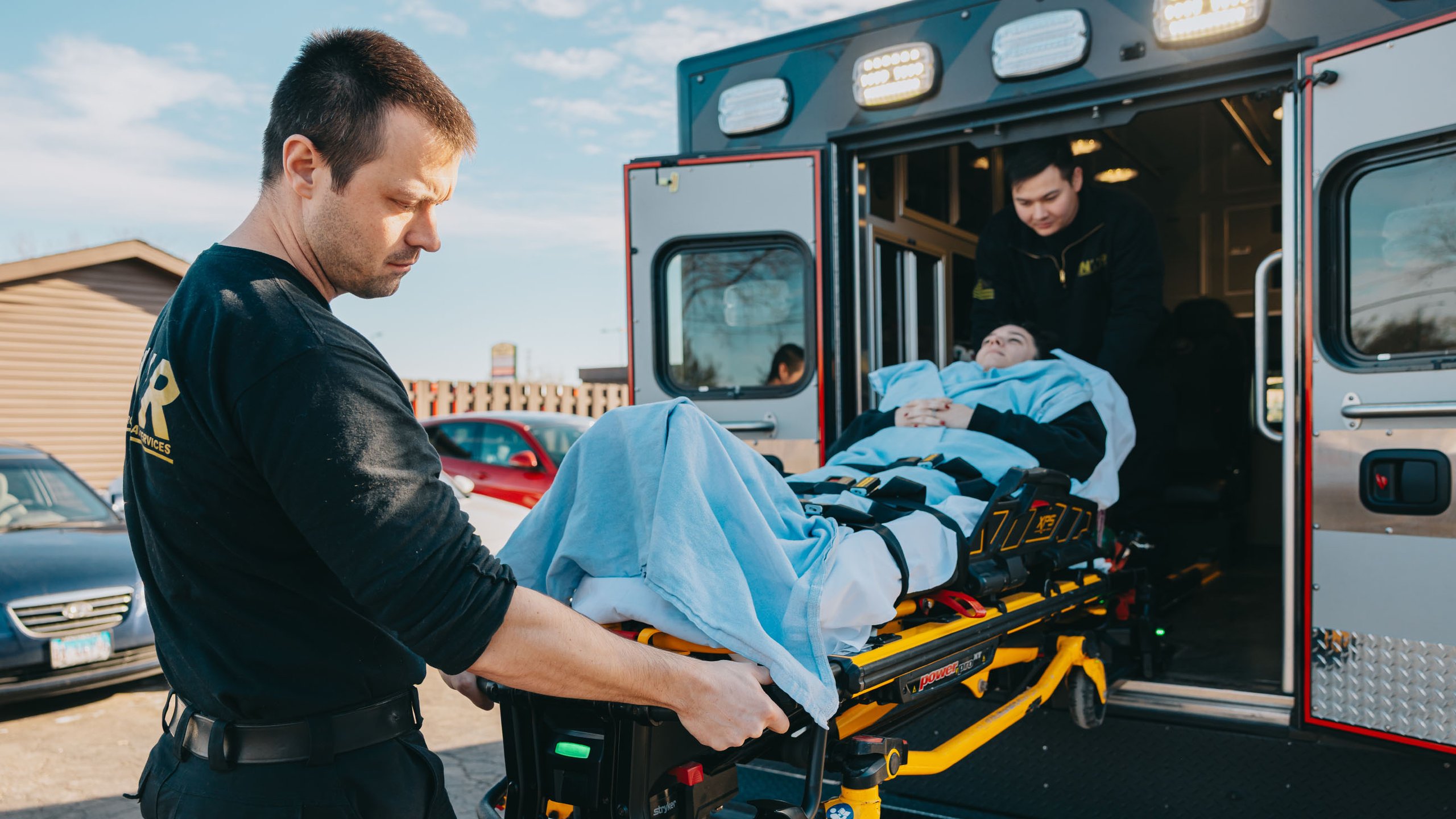 Two paramedics loading a patient covered with a blue blanket into an ambulance on a stretcher.