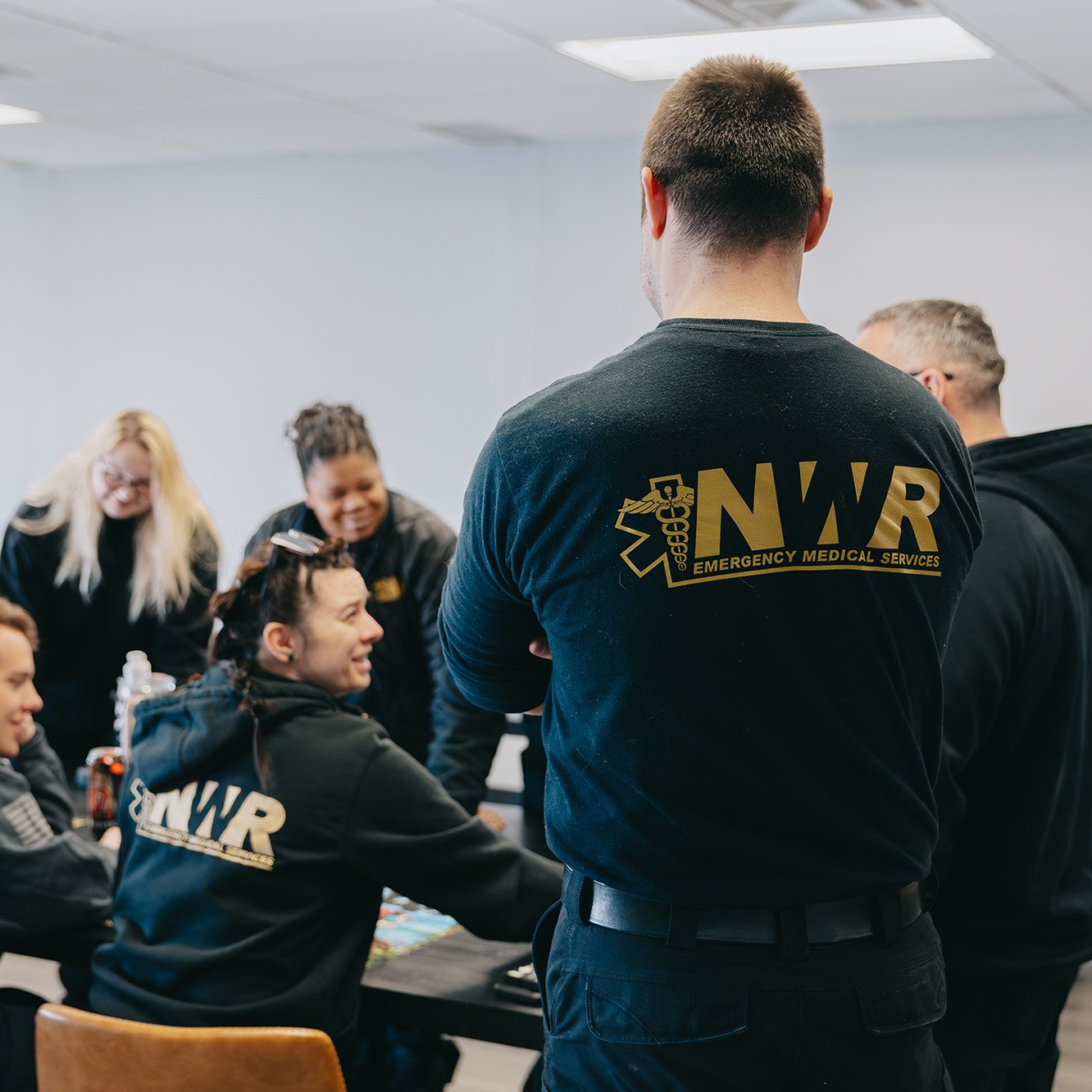 Group of people in black NWR Emergency Medical Services shirts gathered and talking around a table indoors.