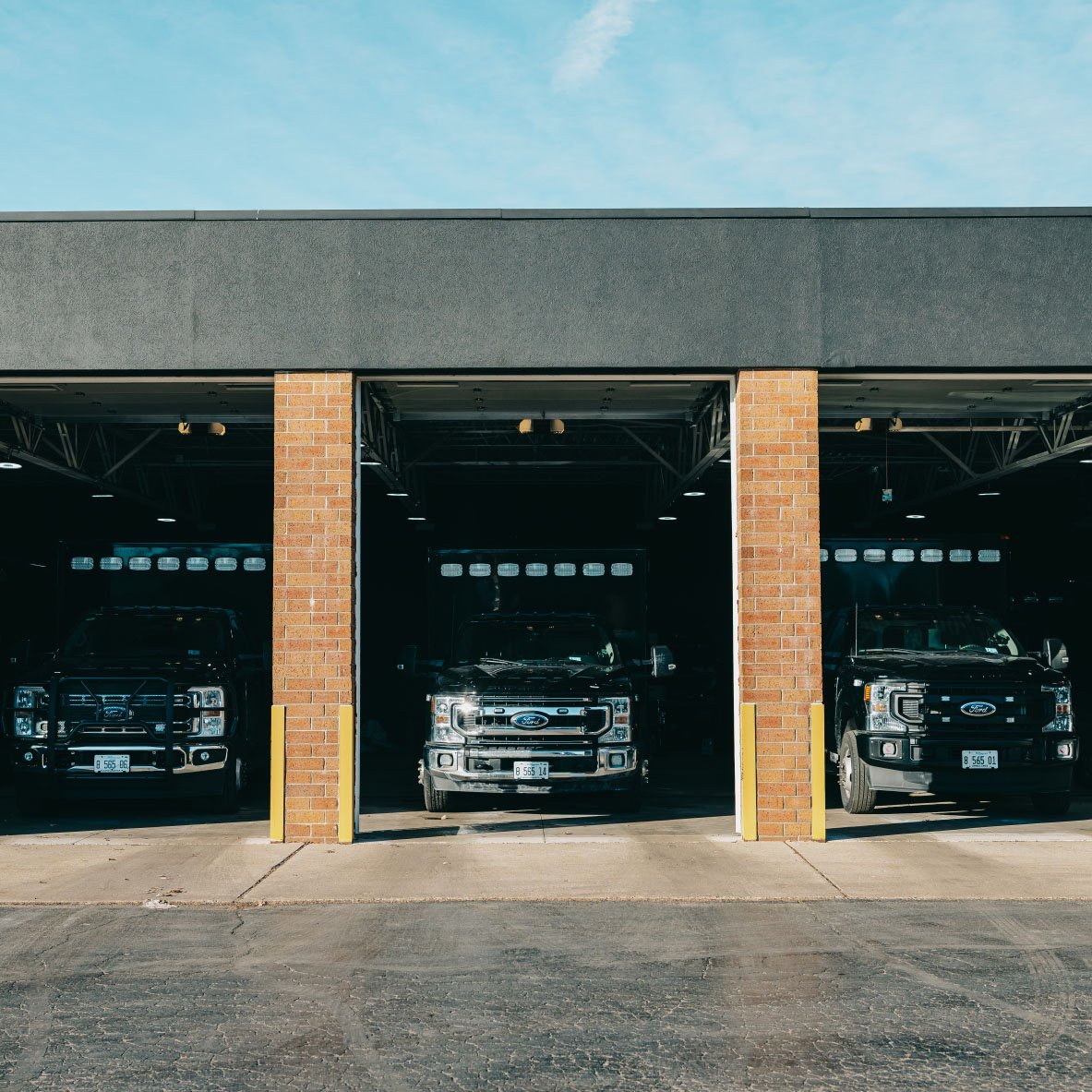Three black Ford trucks parked inside open garage bays with brick pillars under a clear blue sky.