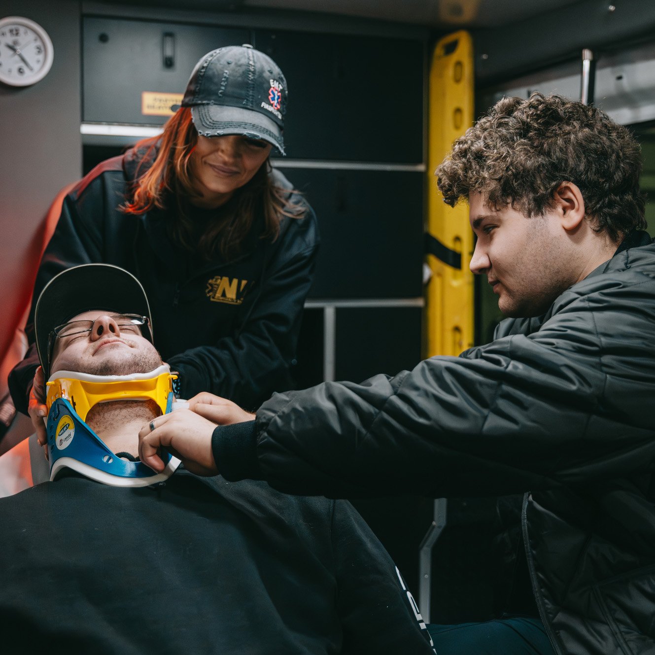 Two paramedics attending to a man lying down with a cervical neck brace inside an ambulance.