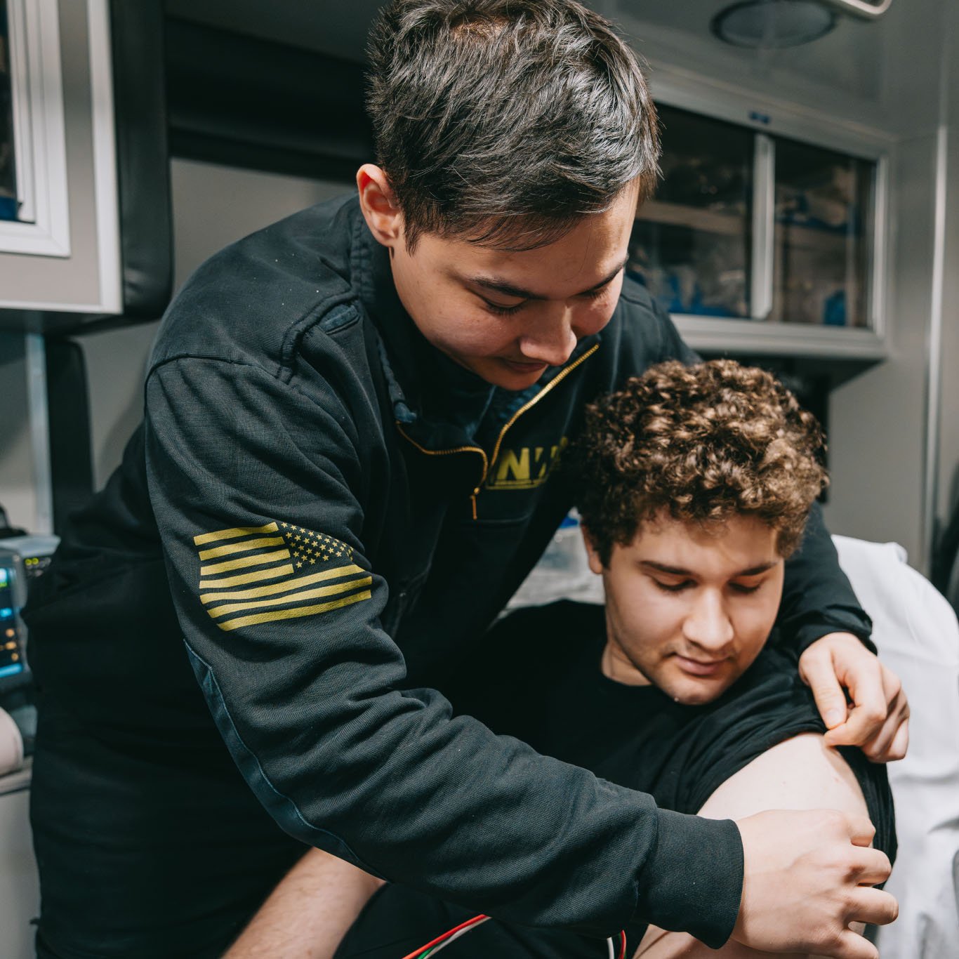 Man in black jacket with American flag patch examining the upper arm of a seated man with curly hair.