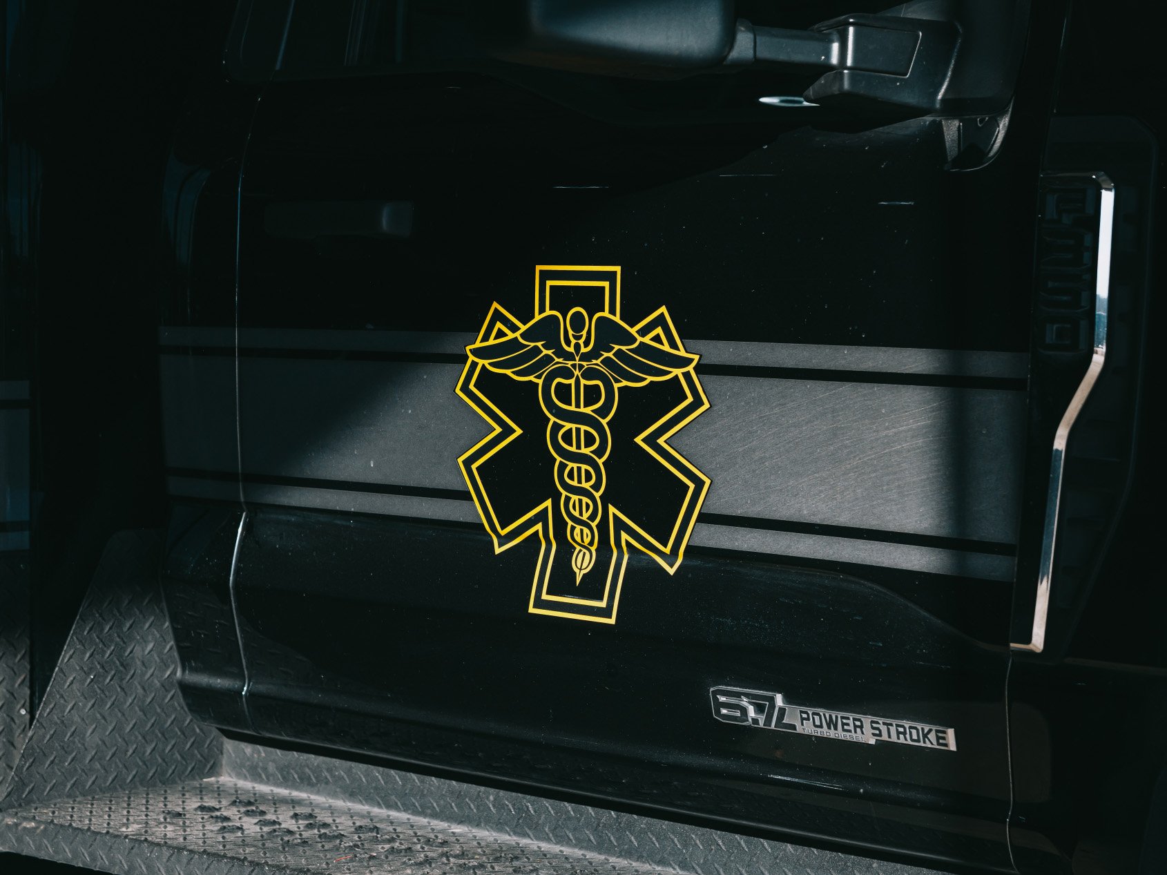 Black vehicle door featuring a yellow medical caduceus symbol and a 6.7L Power Stroke Turbo Diesel badge.