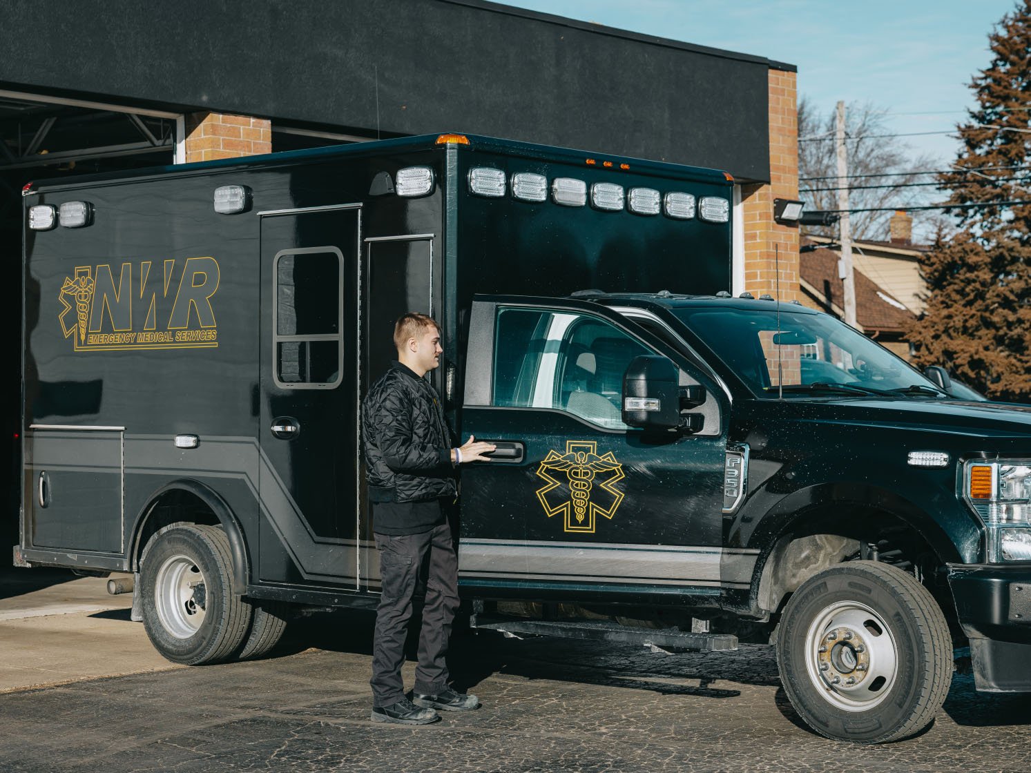 Man standing beside a black NWR Emergency Medical Services ambulance parked outside a brick building.