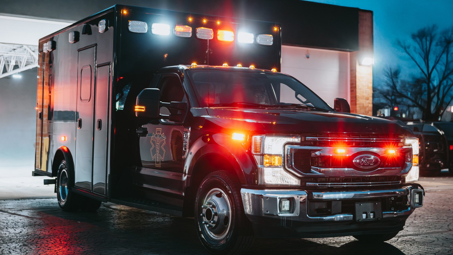 Black Ford ambulance with flashing red and white emergency lights parked outside a building at dusk.