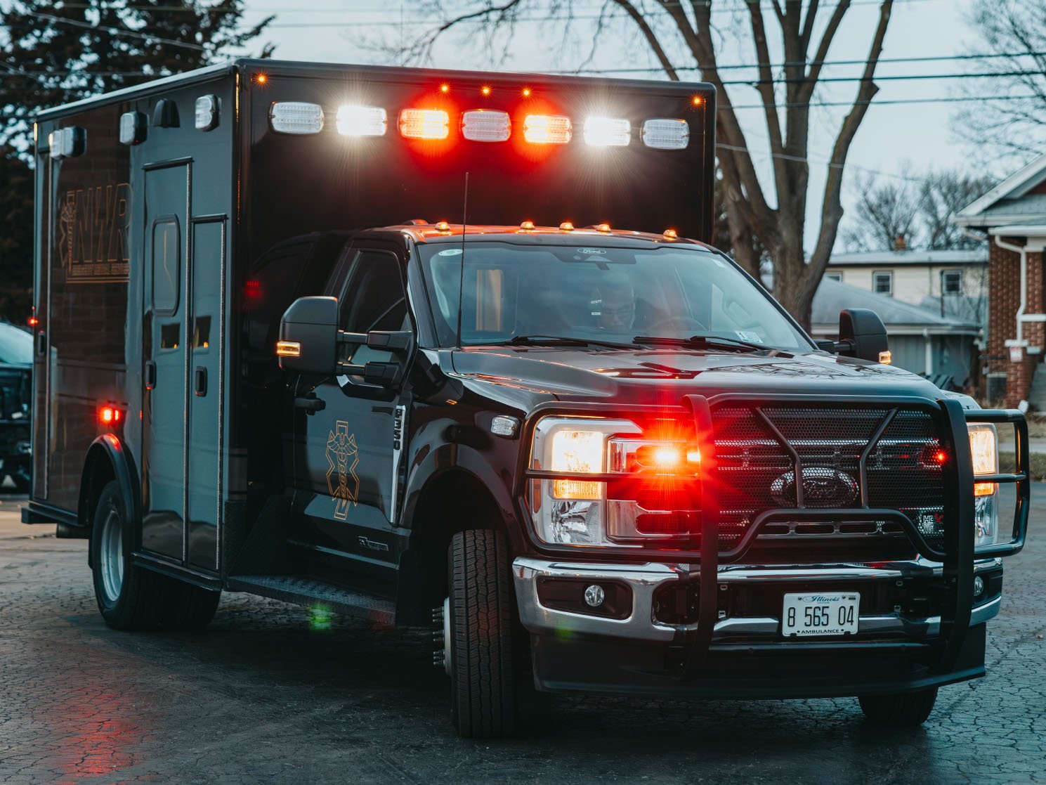 Black emergency ambulance truck with flashing red and white lights parked on a residential street at dusk.