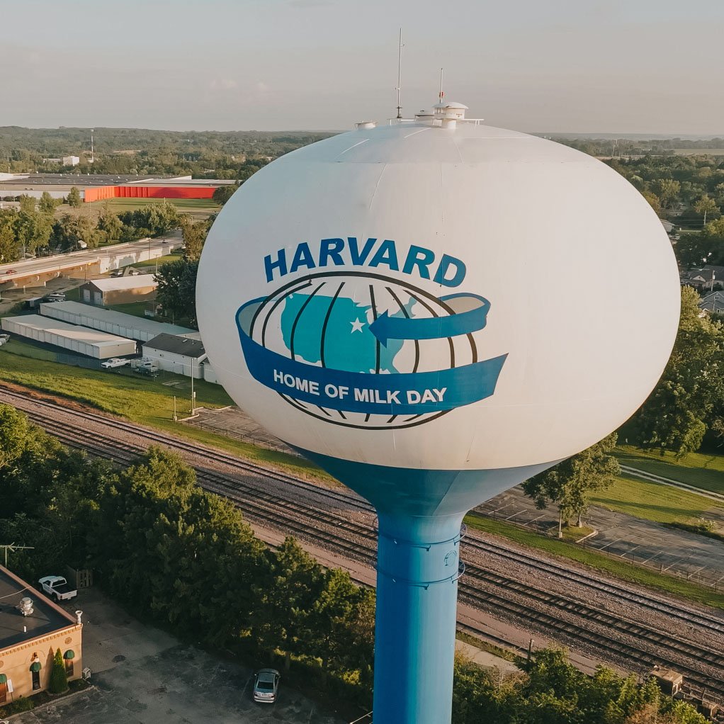 Large white water tower with blue base, displaying a globe and cow silhouette logo with text 'HARVARD HOME OF MILK DAY'.