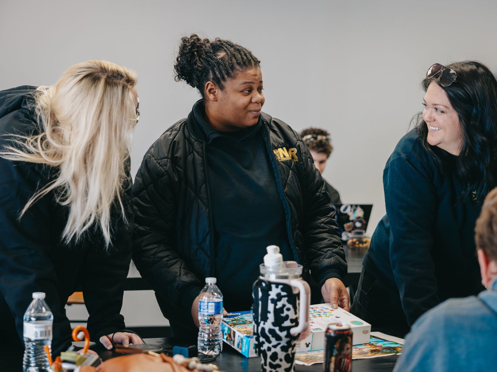 Three women standing around a table engaged in conversation, with puzzle boxes, water bottles, and a drink can on the table.