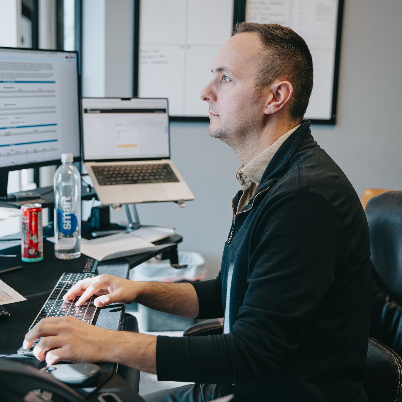 Man focused on typing at a keyboard with dual monitors and a laptop on a desk, drinking water and soda nearby.