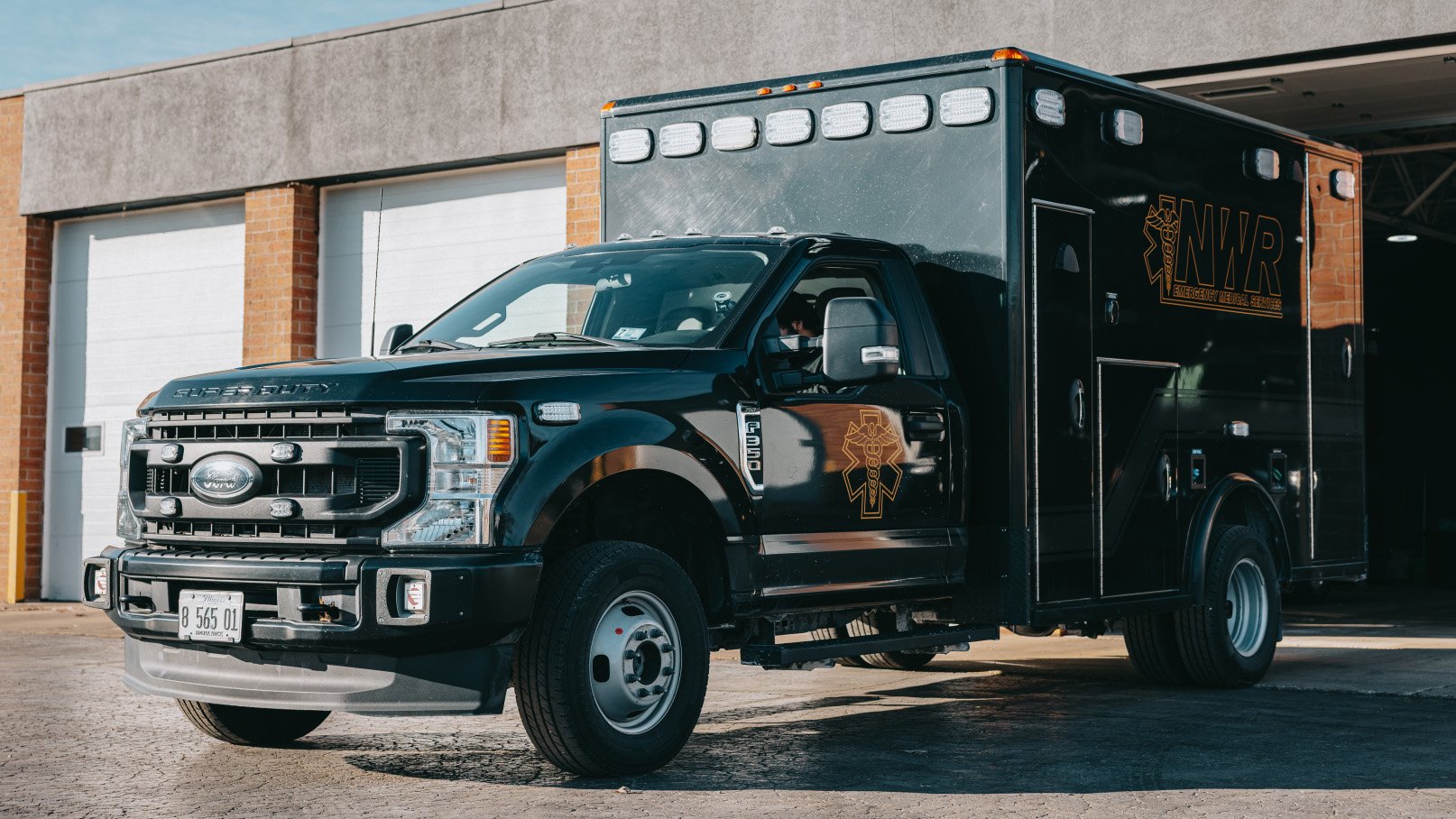 Black Ford Super Duty emergency medical services vehicle parked outside a building with garage doors.